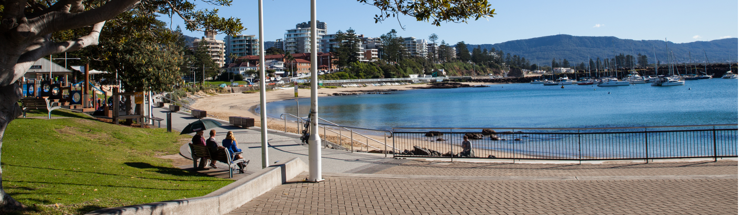 people sitting on a bench on an esplanade overlooking a harbour and mountains