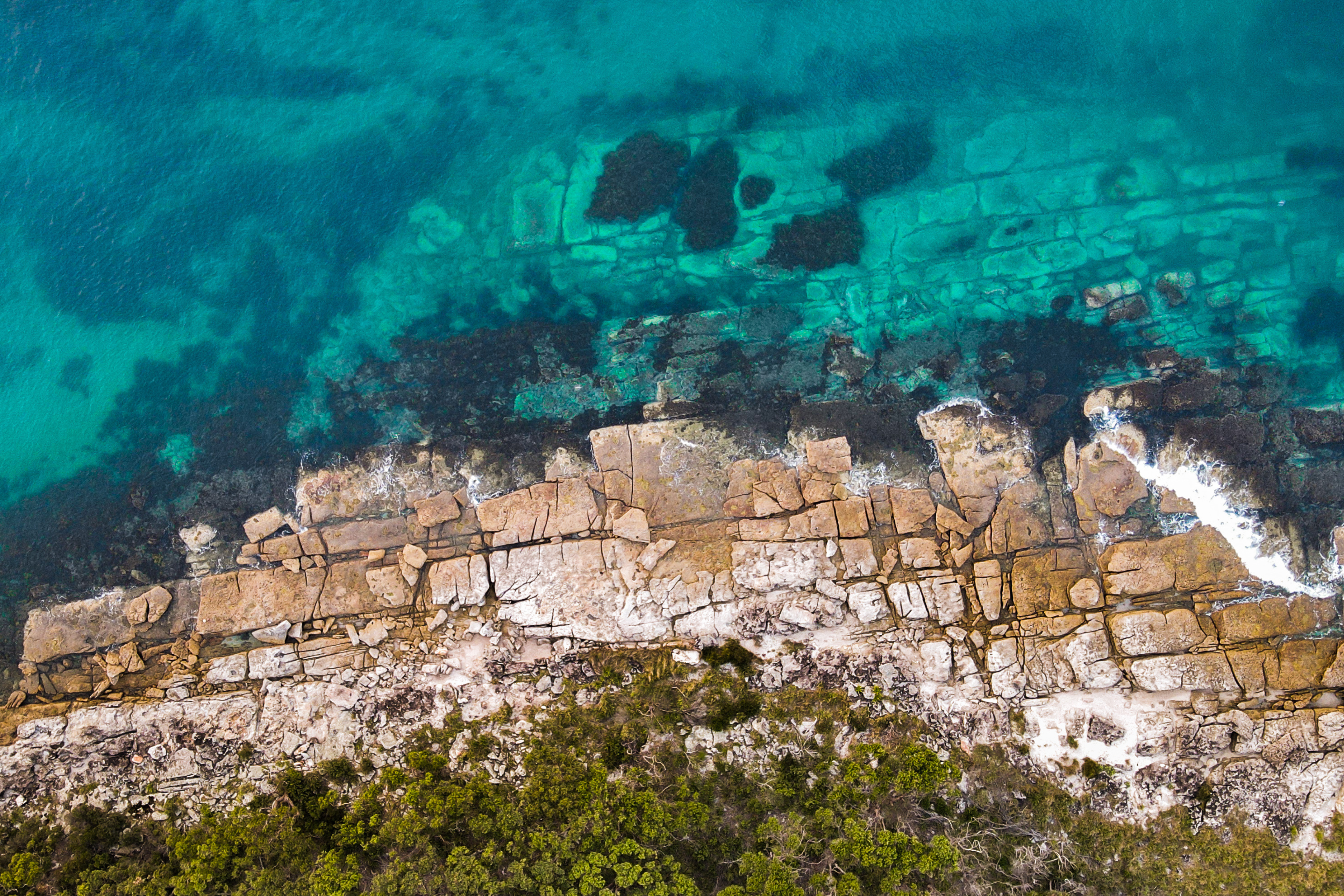 Aerial image of turquoise blue ocean meeting green shrubby land – separated by patterned rocks, at Beecroft Peninsular